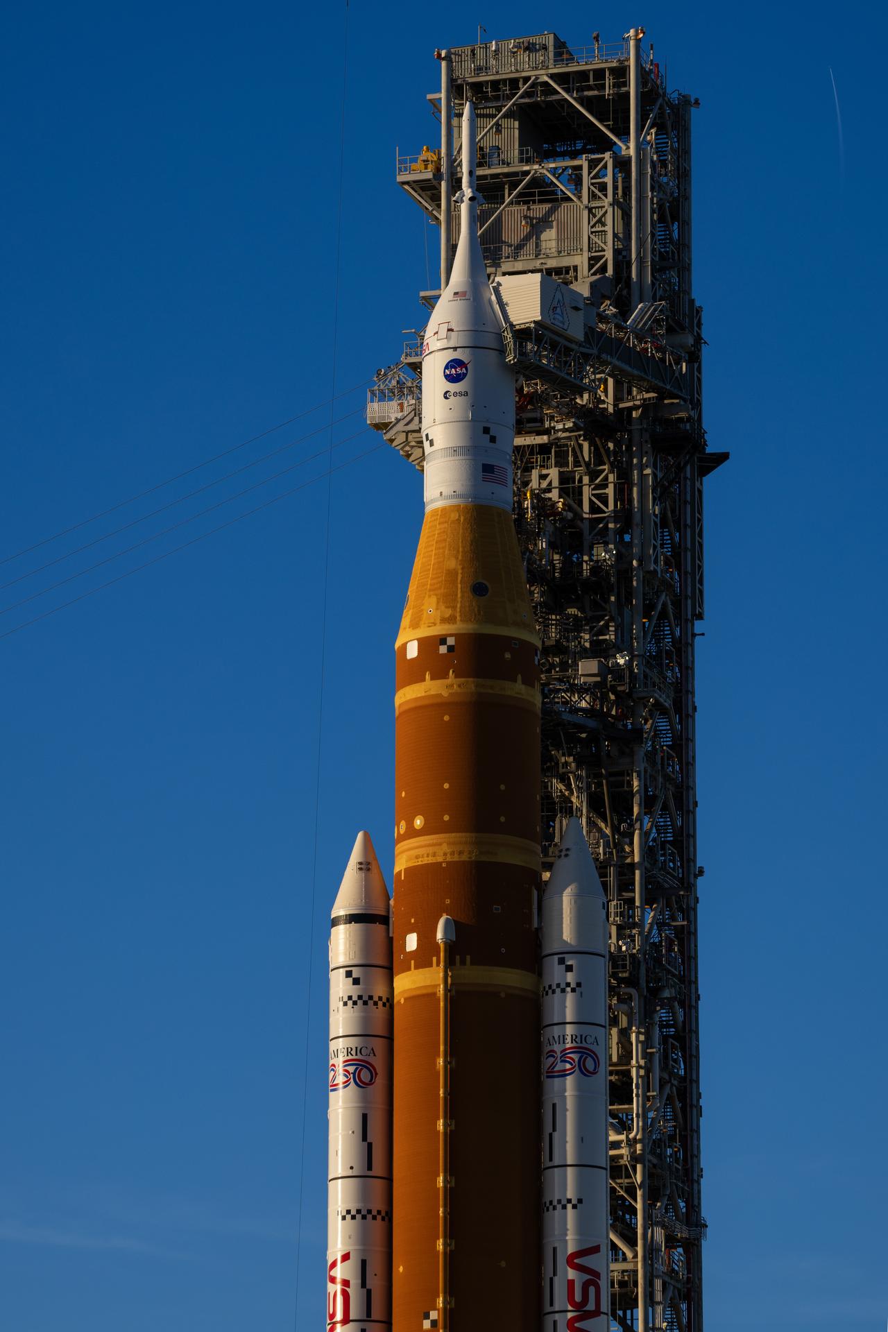 The sun sets on NASA’s Space Launch System (SLS) rocket and Orion spacecraft as they stand fully assembled atop the mobile launcher at Launch Pad 39B at NASA’s Kennedy Space Center in Florida. The sky glows with warm shades of orange and pink, silhouetting the towering rocket and its solid rocket boosters against the fading light. Photographed on January 31, 2026, the scene captures teams preparing for a wet dress rehearsal for the Artemis II mission, rehearsing launch countdown timelines and procedures as day turns to night.
